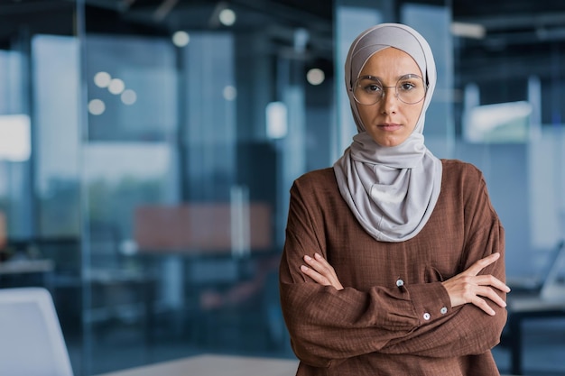 Donna d'affari seria e concentrata in hijab e occhiali che guarda l'obbiettivo con le braccia incrociate che lavorano