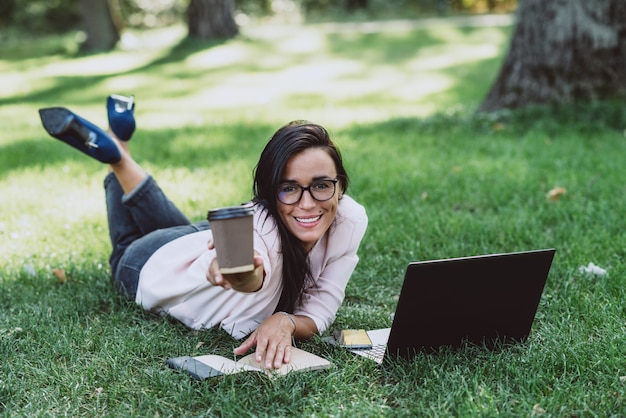 Donna d'affari, si trova in un parco erboso estivo, utilizzando un laptop sorride e porge un bicchiere con il caffè.