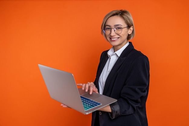 Donna d'affari sicura che indossa un abito nero di stile ufficiale che lavora su un portatile in ufficio guardando la telecamera isolata sullo sfondo arancione