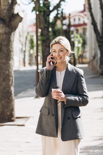 Donna d'affari sorridente bionda matura che cammina per la città bevendo caffè e parlando al telefono in primavera su uno sfondo di fiori lilla