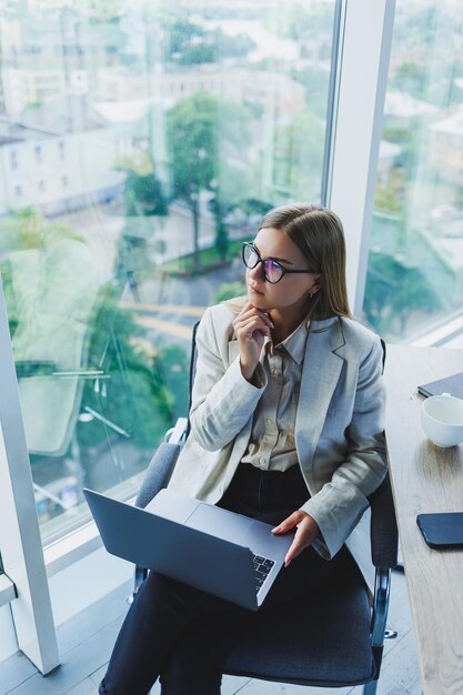 Donna d'affari sorridente che guarda il laptop mentre lavora in ufficio Il concetto di una donna moderna di successo L'idea di affari e la vita di un imprenditore Giovane donna al tavolo con il laptop aperto