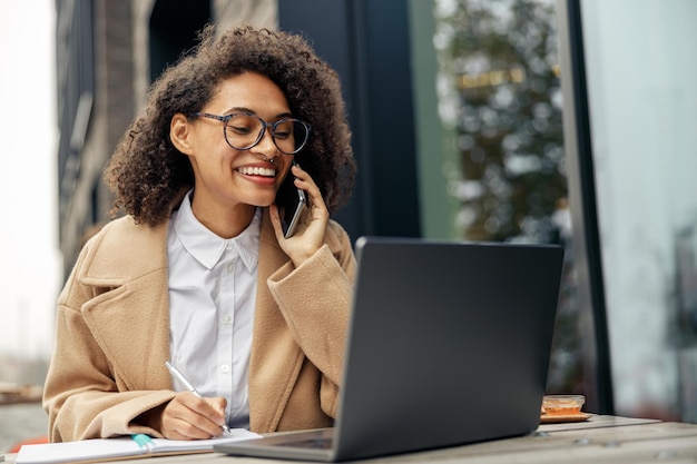 Donna d'affari sorridente che parla al telefono mentre lavora online seduto nella terrazza del caffè