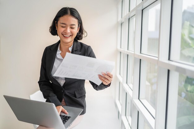 Donna d'affari sorridente con carta in mano e utilizzando il computer portatile in ufficio