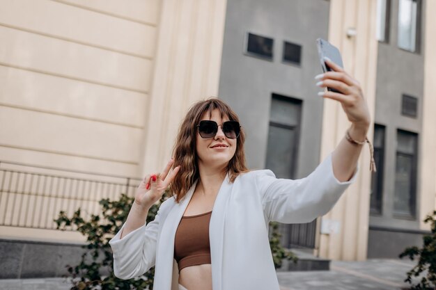 Donna d'affari sorridente in abito bianco che fa videochiamate agli amici durante le passeggiate in città con architettura moderna