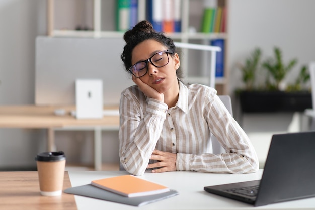 Donna d'affari stanca che dorme sul posto di lavoro mentre lavora al computer portatile nell'interiore moderno dell'ufficio