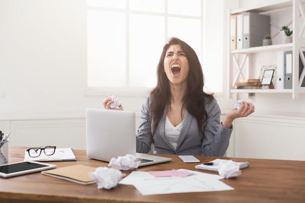 Donna d'affari urlante arrabbiata che lavora con il computer portatile alla scrivania dell'ufficio, copia spazio