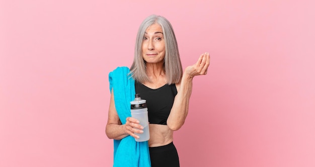 Donna dai capelli bianchi di mezza età che fa un gesto di capice o denaro, dicendoti di pagare con un asciugamano e una bottiglia d'acqua. concetto di fitness