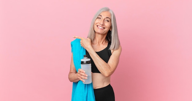 Donna dai capelli bianchi di mezza età che sorride allegramente, si sente felice e indica il lato con un asciugamano e una bottiglia d'acqua. concetto di fitness
