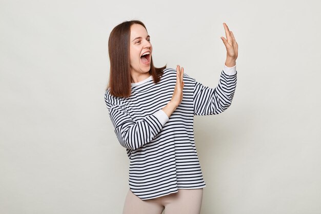 Donna dai capelli castani ottimista positiva che indossa camicia a righe in posa isolata su sfondo grigio in piedi con i palmi sollevati urlando di felicità umore festivo