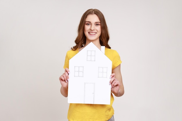 Donna dai capelli castani positiva di giovane età con capelli ondulati in maglietta gialla che tiene una casa di carta con un'espressione felice, sognando la propria casa. Studio indoor girato isolato su sfondo grigio.