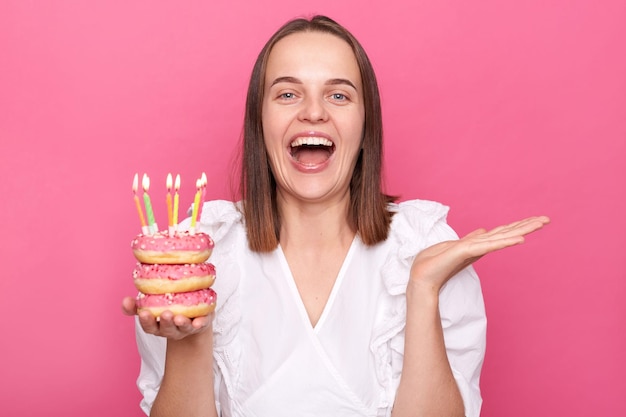 Donna dai capelli castani soddisfatta che tiene deliziose ciambelle appetitose con candele accese indossa camicetta bianca che festeggia il compleanno da sola a casa isolata su sfondo rosa