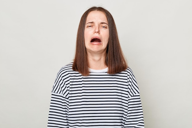 Donna dai capelli castani triste e infelice che indossa una camicia a righe in posa isolata su sfondo grigio piangendo sensazione di disperazione sentendo notizie molto cattive