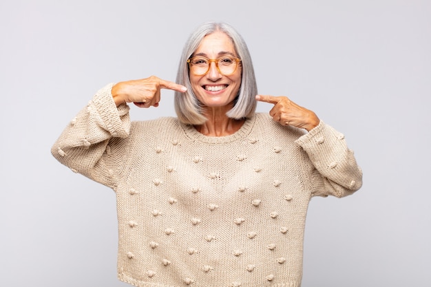 Donna dai capelli grigi che sorride fiduciosamente indicando il proprio ampio sorriso, atteggiamento positivo, rilassato, soddisfatto