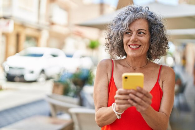 Donna dai capelli grigi di mezza età che sorride fiduciosa utilizzando lo smartphone in strada