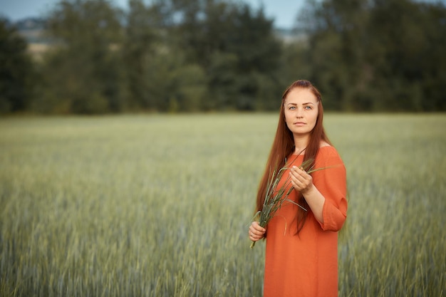 Donna dai capelli rossi caucasica in un vestito rosso che cammina su un campo di fattoria con grano al tramonto su un raccolto di cibo biologico estivo dayfuturo