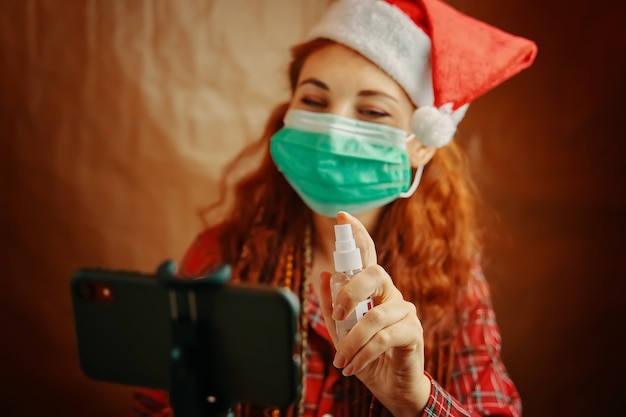 Donna dai capelli rossi con cappello da Babbo Natale e maschera medica spruzza antisettico sul suo smartphone natale a quara...