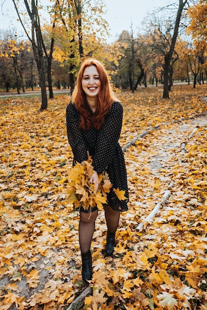 Donna dai capelli rossi felice che tiene le foglie di autunno dell'acero giallo nel parco di caduta