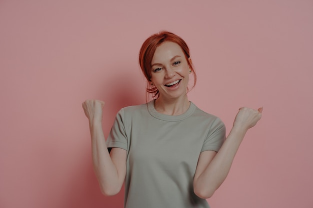 Donna dai capelli rossi felicissima che celebra la vittoria facendo il gesto del vincitore su sfondo rosa