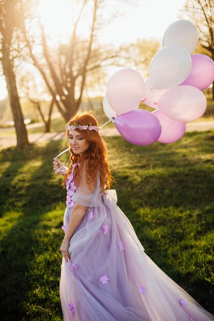 Donna dai capelli rossi in abito viola a piedi nel parco con palloncini in mano, al tramonto