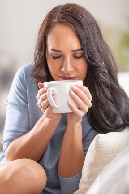Donna dai capelli scuri che si gode una bella tazza di tè o caffè mentre è seduta su un divano.