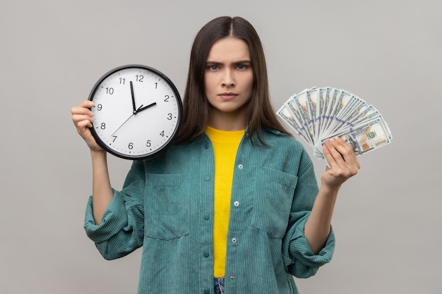 Donna dai capelli scuri ricca e seria che tiene in mano un grande fan delle banconote in dollari e il tempo dell'orologio da parete è denaro