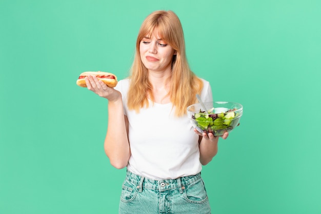 Donna dalla testa abbastanza rossa che tiene un'insalata e un hot dog. concetto di dieta