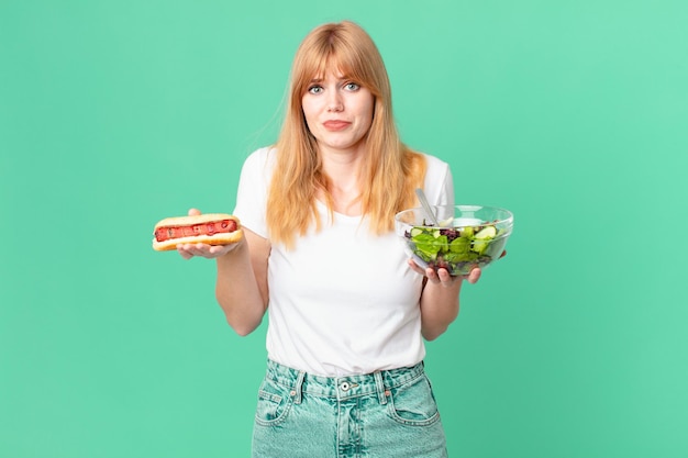 Donna dalla testa abbastanza rossa che tiene un'insalata e un hot dog. concetto di dieta