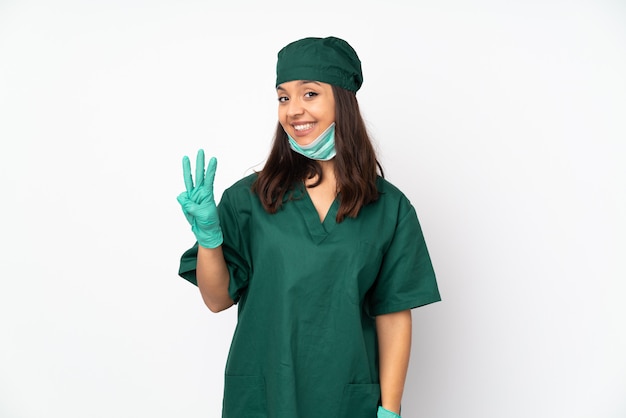 Donna del chirurgo in uniforme verde sulla parete bianca felice e contando tre con le dita