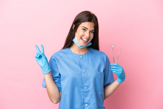 Donna del dentista che tiene gli strumenti isolati su sfondo rosa sorridendo e mostrando il segno della vittoria