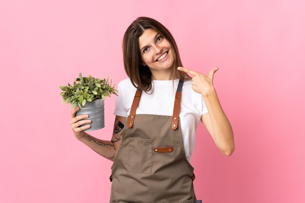 Donna del giardiniere che tiene una pianta isolata sulla parete rosa che dà un pollice in alto gesto