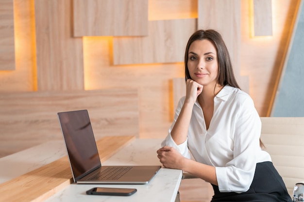 Donna del responsabile dell'ufficio in camicia bianca bel sorriso seduto al tavolo con il portatile mano al mento nell'interno dell'ufficio in legno Impiegato amichevole che esamina la macchina fotografica Concetto di lavoro