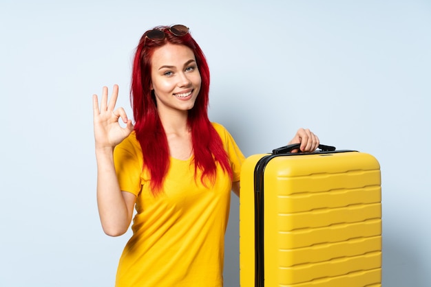 Donna del viaggiatore che giudica una valigia isolata sulla parete blu che mostra segno giusto con le dita