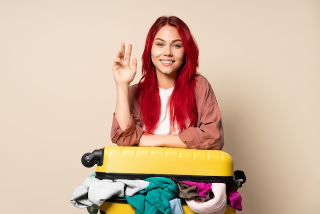 Donna del viaggiatore con una valigia piena di vestiti isolati sulla parete beige che saluta con la mano con l'espressione felice