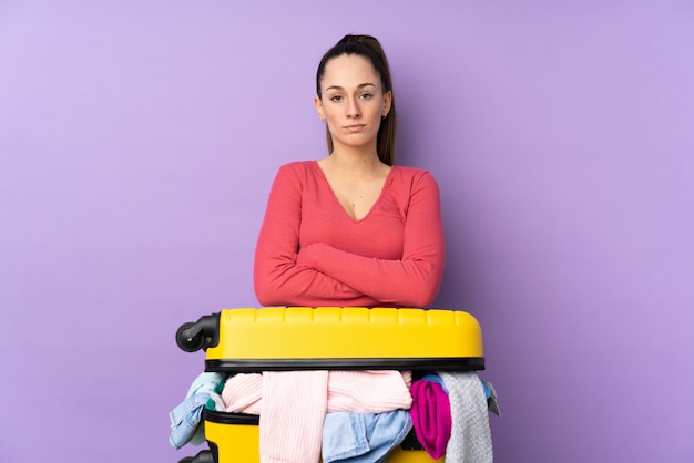 Donna del viaggiatore con una valigia piena di vestiti sopra il muro viola isolato mantenendo le braccia incrociate