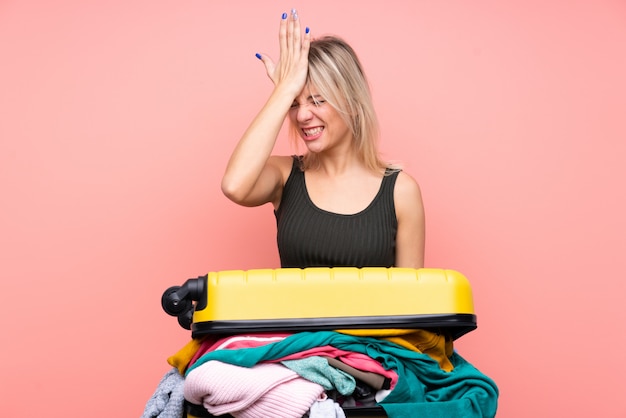 Donna del viaggiatore con una valigia piena di vestiti sopra la parete rosa isolata che ha dubbi con l'espressione del viso confuso