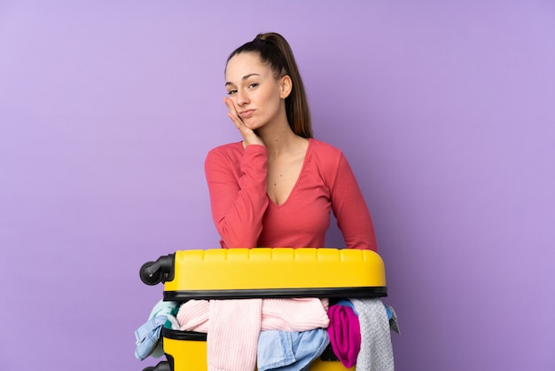 Donna del viaggiatore con una valigia piena di vestiti sopra la parete viola isolata infelice e frustrata