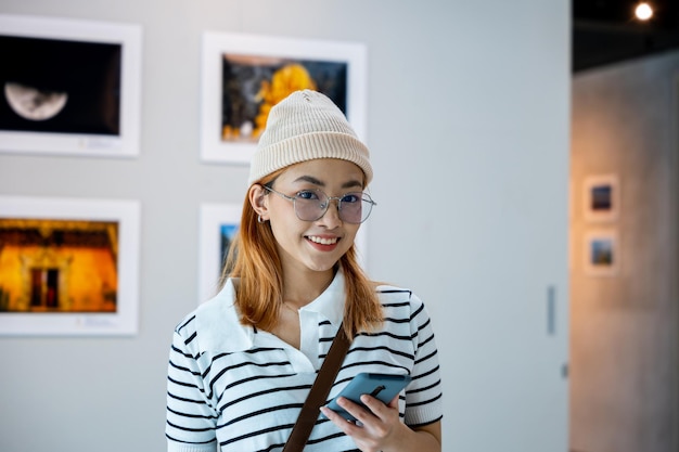 Donna del visitatore che sorride alla collezione della galleria d'arte nella parte anteriore dei dipinti incorniciati che tiene il telefono cellulare