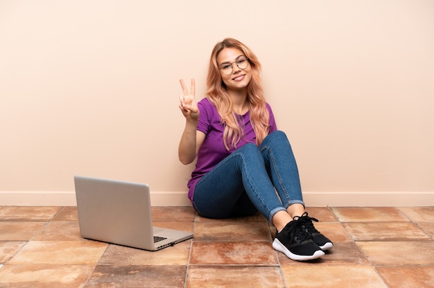 Donna dell'adolescente con un computer portatile che si siede sul pavimento a sorridere al chiuso e che mostra il segno di vittoria