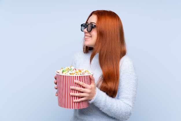 Donna dell'adolescente della testarossa sopra la parete blu isolata con gli occhiali 3d e tenere un grande secchio di popcorn