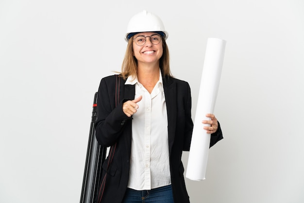 Donna dell'architetto di mezza età con il casco e che tiene i modelli sopra la parete isolata sorpresa e che indica davanti