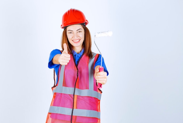 Donna dell'ingegnere in uniforme che tiene un rullo di pittura colorato bianco e che mostra il segno di soddisfazione.