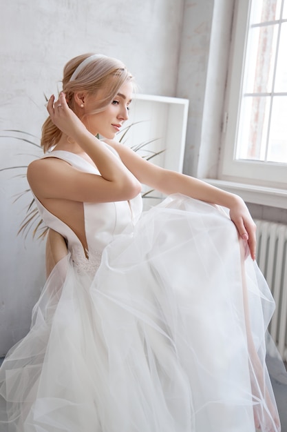 Donna della sposa in un vestito da sposa leggero da estate che si siede alla finestra e che aspetta la cerimonia di nozze. Ragazza con capelli perfetti e bellissimo trucco