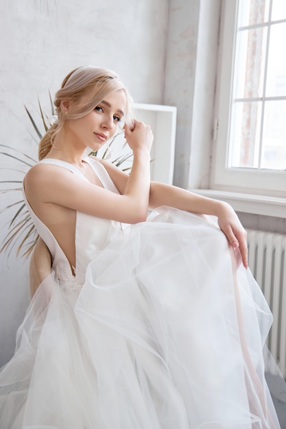 Donna della sposa in un vestito da sposa leggero da estate che si siede alla finestra e che aspetta la cerimonia di nozze.