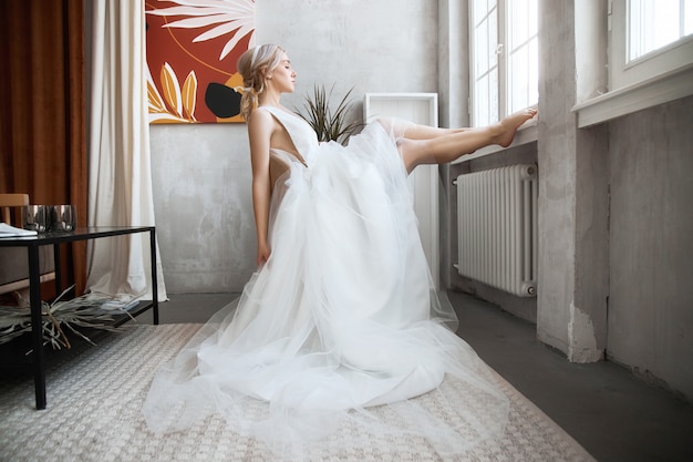 Donna della sposa in un vestito da sposa leggero da estate che si siede alla finestra e che aspetta la cerimonia di nozze