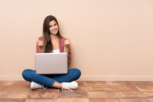 Donna dello studente dell'adolescente che si siede sul pavimento con un computer portatile che fa gesto dei soldi