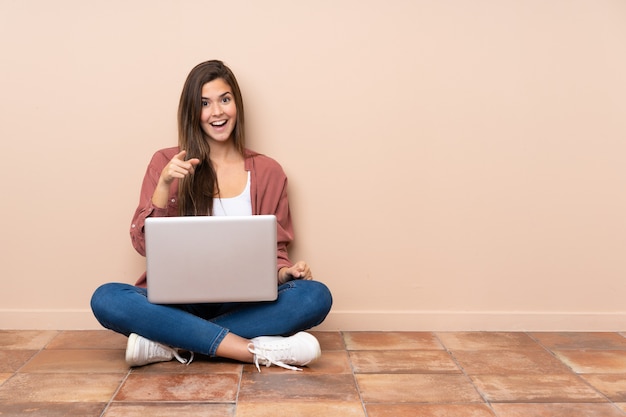 Donna dello studente dell'adolescente che si siede sul pavimento con un computer portatile sorpreso e che indica parte anteriore