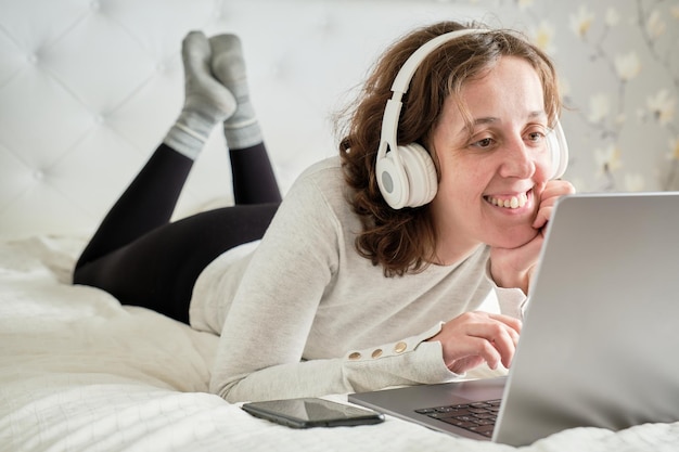 Donna di 45 anni con le cuffie che ascolta la musica sul suo laptop sdraiato sul letto