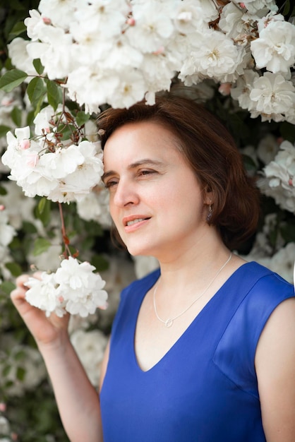 Donna di 48 anni con un vestito blu vicino a un cespuglio di rose bianche in fiore