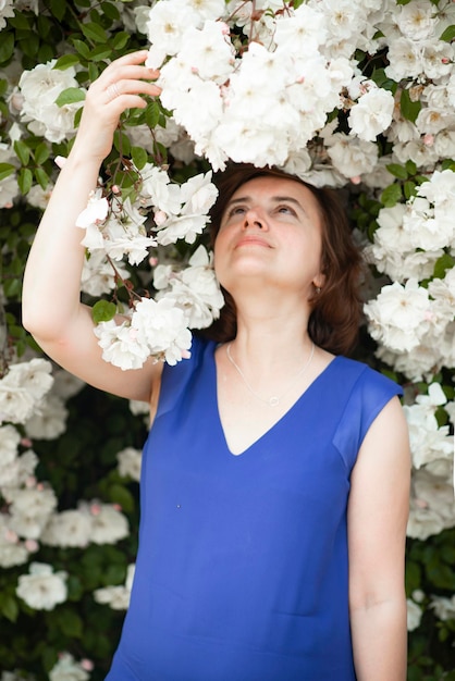 Donna di 48 anni in un vestito blu vicino a un cespuglio di rose bianche in fiore