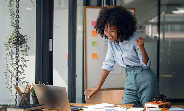 Donna di affari afroamericana di successo ed emozionante entusiasta di vincere sul suo computer portatile in piedi in ufficio